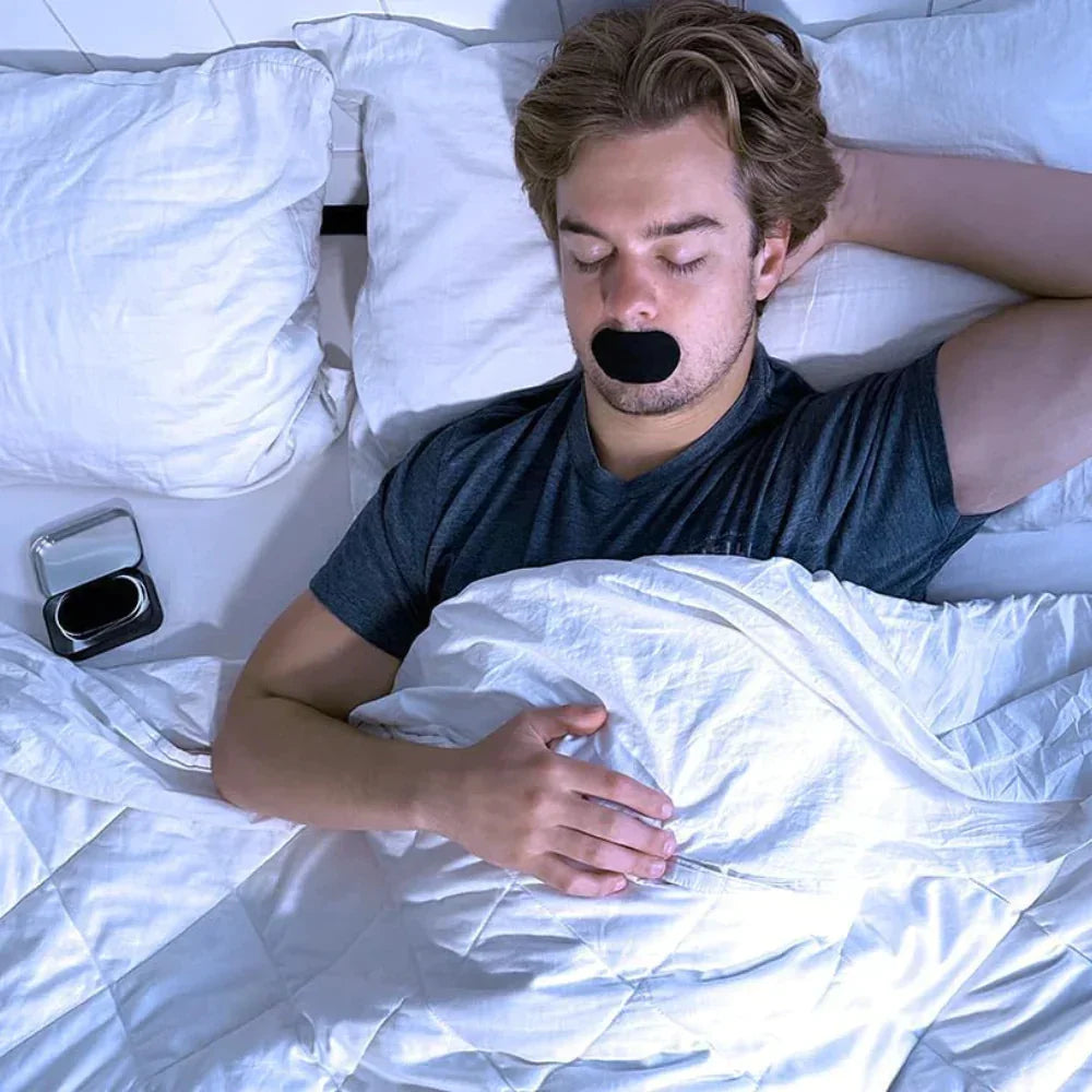 Man sleeping on white bed with black anti-snoring mouth patch applied over lips.